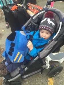 A toddler riding in a gray stroller hugging his stuffed Popatu blue airplane wheeled suitcase