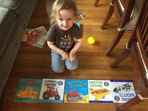 Toddler with five titles from Amazing Machines paperback book series by Tony Mitton laid out on floor