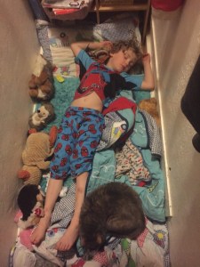 Child asleep on closet floor surrounded by stuffed animals and blankets