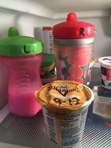 Two Playtex sippy cups and an open container of yogurt labeled with dry erase marker on top shelf of fridge