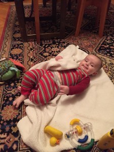 Infant in red striped pajamas lying on floor on top of towel with toys around