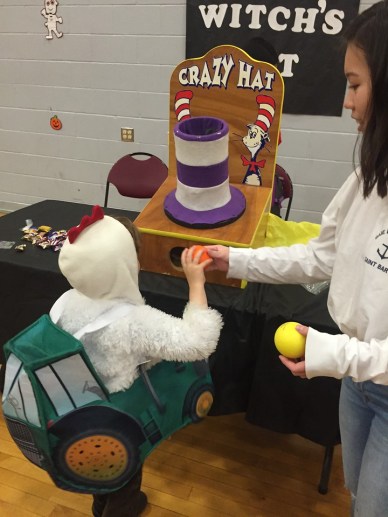 Child in chicken tractor costume throwing balls into tall striped hat carnival game