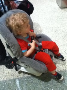 Toddler riding in car seat on travel cart wheels in airport next to suitcase