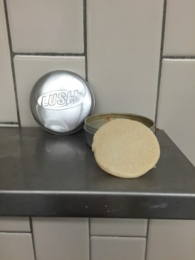 Lush solid shampoo in Godiva scent sitting on shower shelf propped up on tin