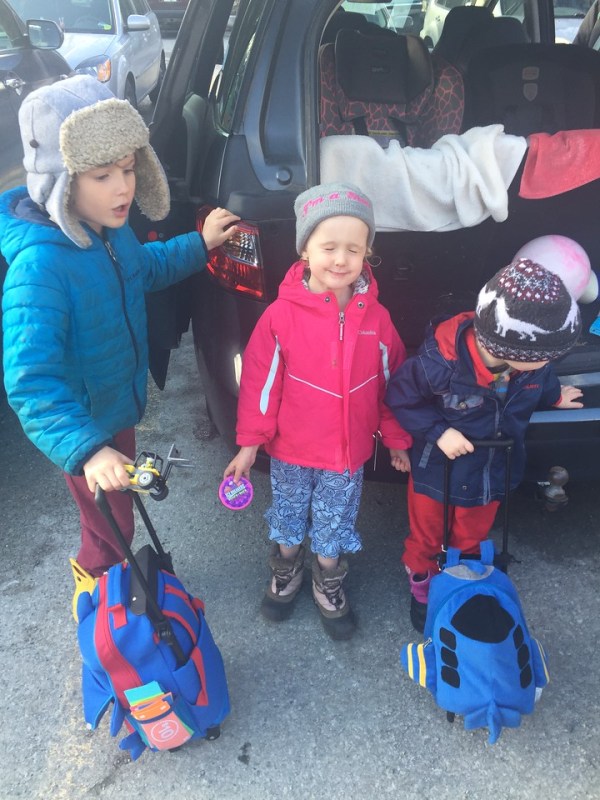 Three kids with rolling backpack suitcases wearing snow boots for travel