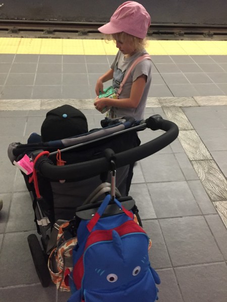 Kid standing next to child in stroller loaded down with bags on train platform