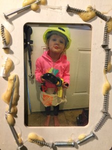 Child wearing hard hat and holding drill in photo frame decorated with nails and screws