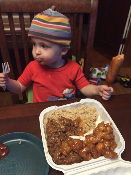 Toddler with corn dog in one hand and fork in other in front of food at table