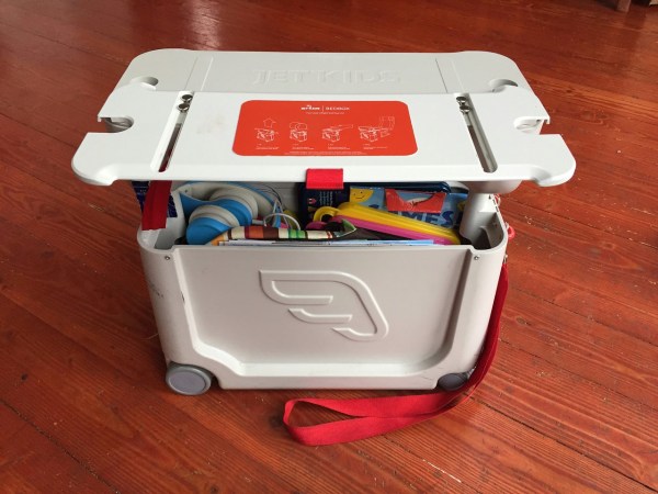 JetKids BedBox packed with travel supplies inside shown in bed mode with platform open on top of ride on suitcase