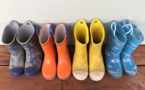 Croc Bump It and Norty kids rain boots four pairs in a row on a wooden porch floor shark print, orange and blue, bright yellow, and blue unicorn print