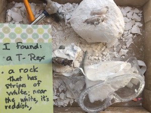 Dinosaur dig with rock inside plaster with small hammer, kid sized safety googles, brush, and list of what was found inside