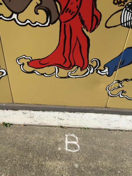 Letter B drawn in sidewalk chalk on concrete below art on building