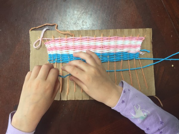 Child weaving pink white and blue project on homemade cardboard loom