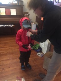 Child getting ready to leave house wearing pink rain jacket, black rain pants, waterproof boots, hat, gloves, and face mask