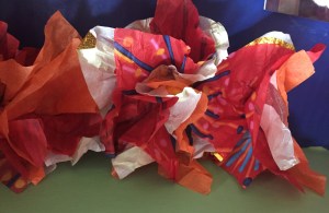 Three tissue paper flowers in a row made from striped white paper, striped red paper, and orange paper by eight year old