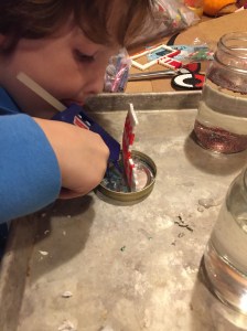 Child using hot glue gun to attach metly bead ornament to inside of jar lid on a cookie sheet