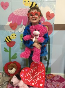Preschooler child wearing heart glasses frames hugging pink teddy bear with heart flowers in background and Happy Valentine's Day sign