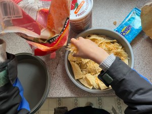 Kid in kitchen assembling their own plate of nachos with tortilla chips and cheese in cake layer pan
