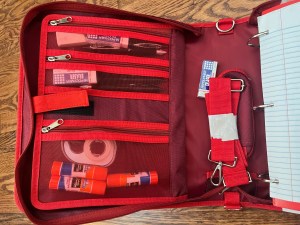Red three ring binder with zipper and carrying strap included shown holding glue sticks, erasers, pens, and other school supplies in organizer