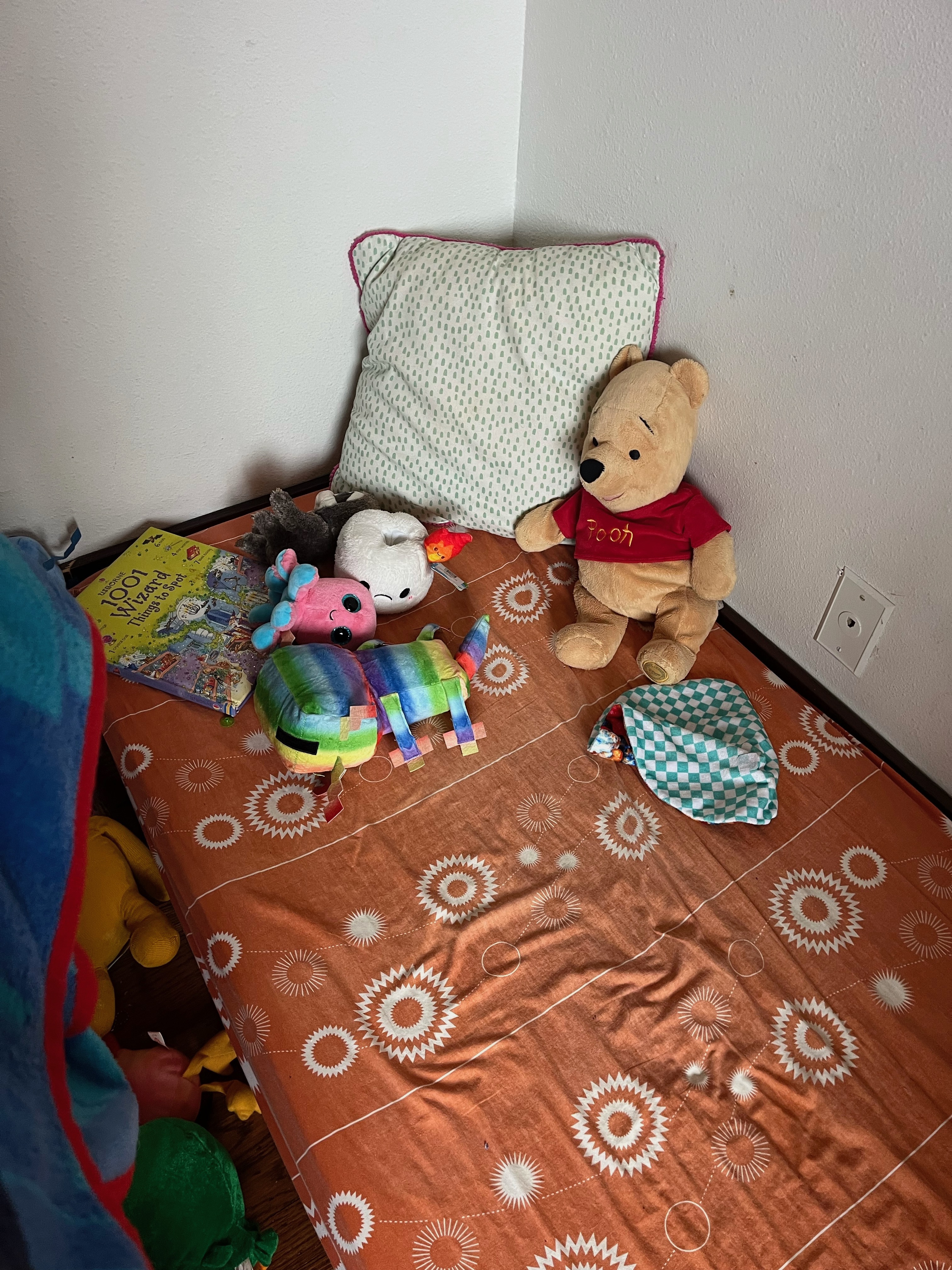 Crib mattress with orange sheet, pillow, and Pooh Bear stuffed animal on floor in kid's bedroom