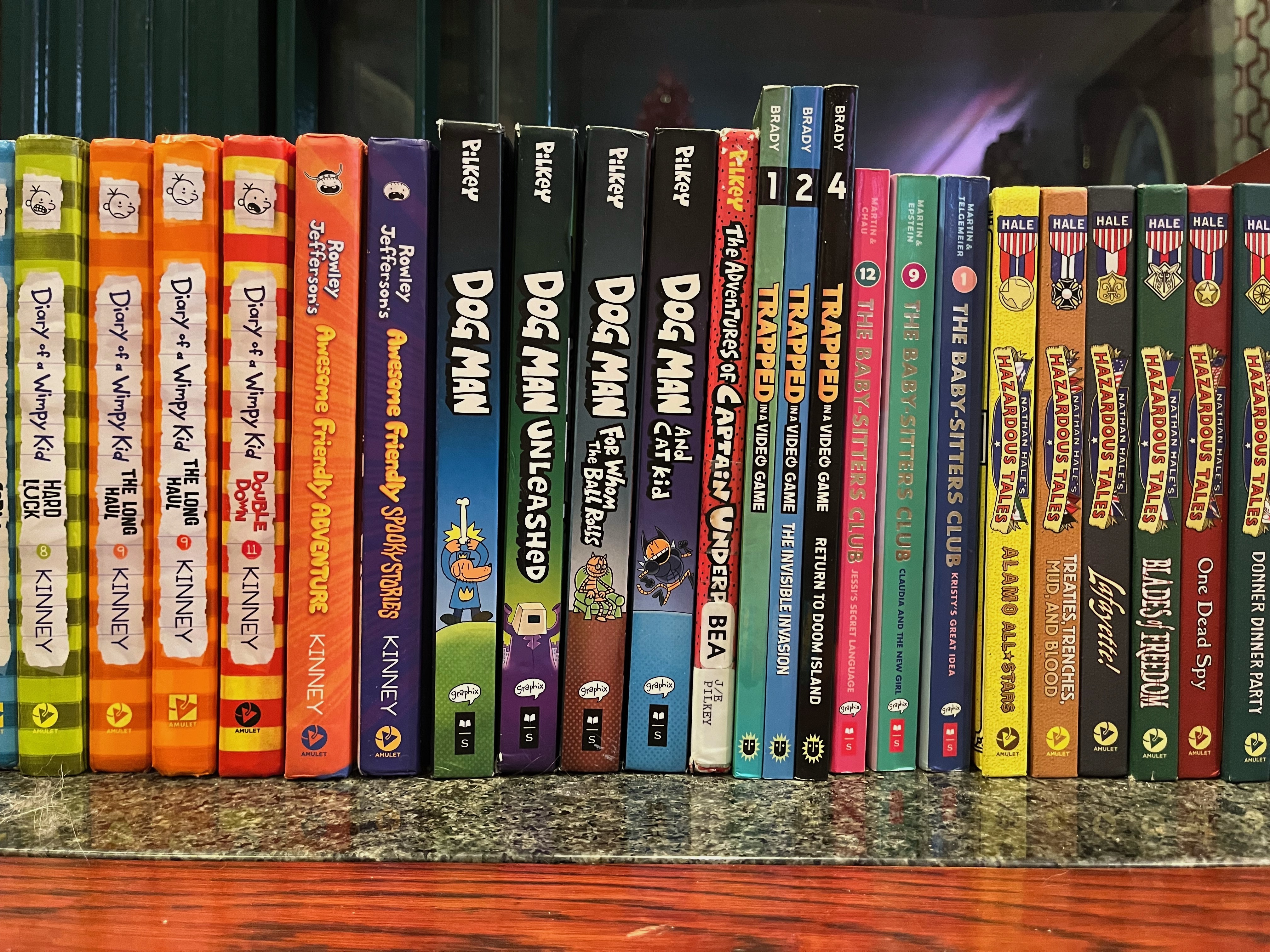 Book series for kids lined up on fireplace mantel Diary of a Wimpy Kid, Dog Man, Captain Underpants, Trapped in a Video Game, The Baby-Sitters Club, Nathan Hale's Hazardous Tales